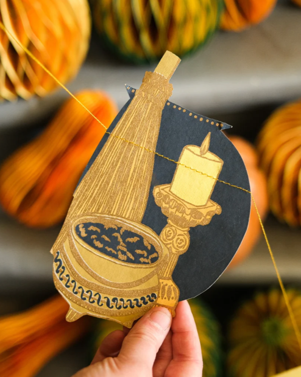Person holding a decorative paper cutout featuring a candle on a candlestick, a vase, and a bowl of fruit, set against a background of striped gourds.