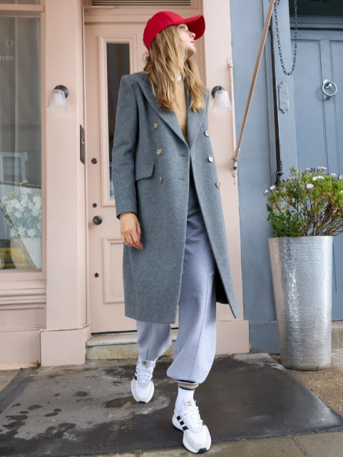 Young woman in a gray coat, red cap, and sweatpants standing outside a storefront with a pink door and large silver planter.