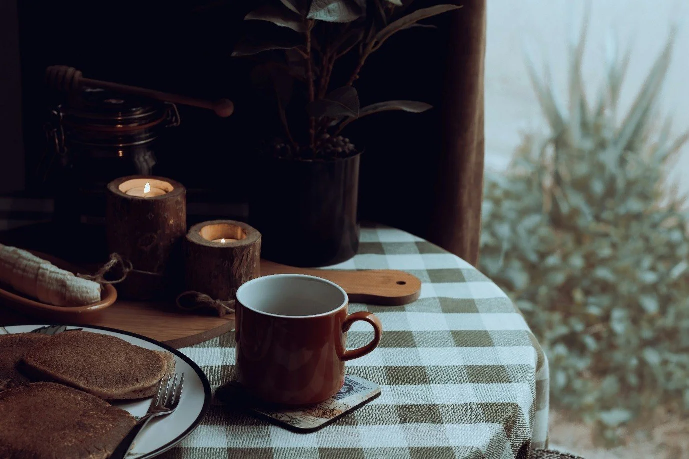 A table with a checkered tablecloth holds a plate of toast, a red mug, two lit candles in wooden holders, and a potted plant, with a window showing outside foliage in the background.