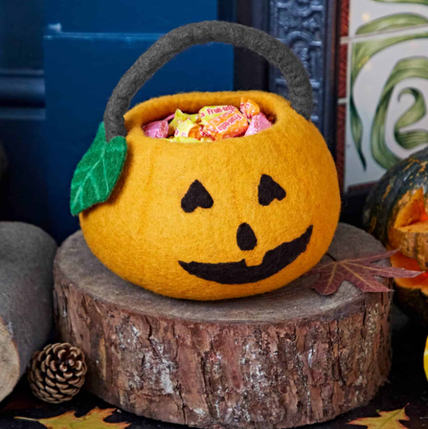 Felt pumpkin-shaped container with a jack-o'-lantern face, filled with Halloween candies, placed on a tree stump surrounded by fall decor.