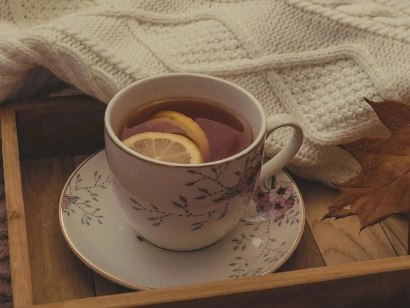 A floral patterned teacup filled with tea and lemon slices, placed on a matching saucer, with a cream-colored knitted blanket and autumn leaf nearby, inside a wooden tray.
