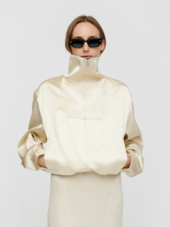 Woman wearing a high-collared cream jacket and black sunglasses against a plain gray background.