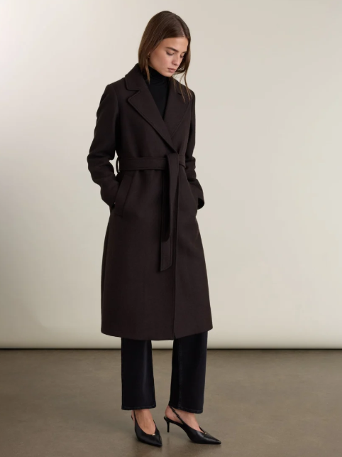 Woman in a black coat, black pants, and black heels standing against a plain beige background.