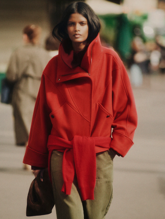Woman walking outdoors wearing a red oversized coat with a matching scarf tied around her waist, holding a bag in her left hand, with blurred people in the background.