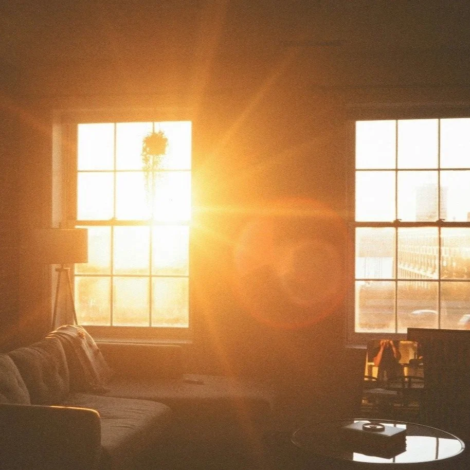 Sunlight streams through two large windows, illuminating a cozy living room with a sofa, a small table, and a lamp, creating a warm, golden glow.