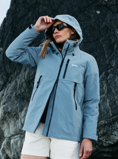 A woman wearing a light blue waterproof jacket and black sunglasses standing outdoors against a rocky background.