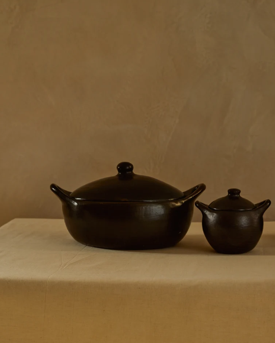 Black ceramic pots with lids, one larger and one smaller, placed on a table with a beige tablecloth against a light-colored wall.