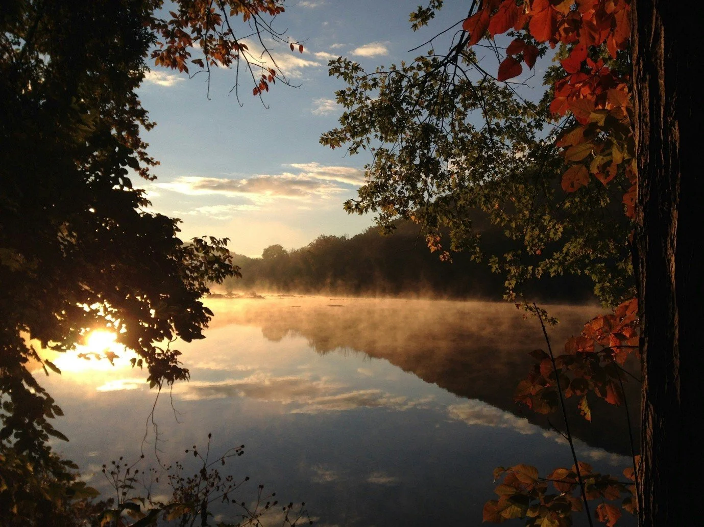 A serene lake surrounded by trees with autumn leaves, fog rising from the water, and a partly cloudy sky at sunrise or sunset.