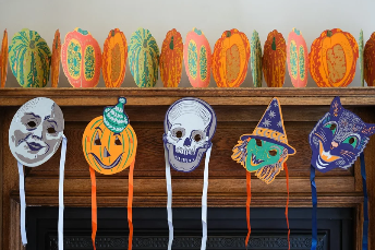 Halloween-themed paper pumpkin decorations on a mantel with masks on sticks in front of them.