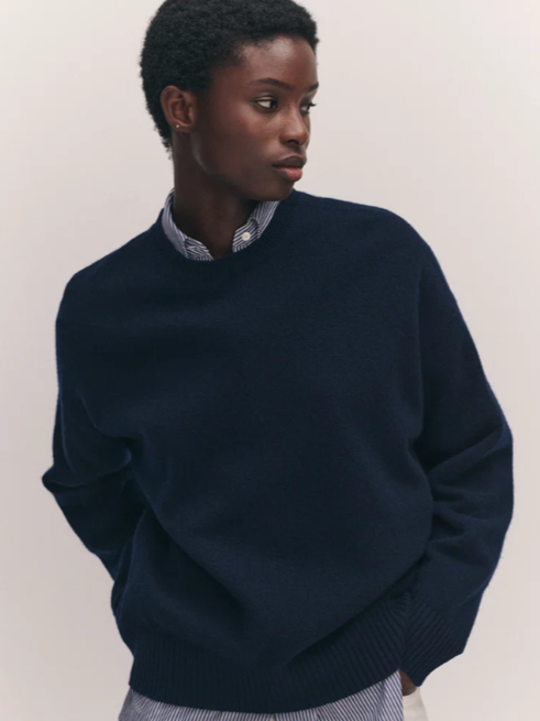 Young woman with short natural hair wearing a dark blue sweater and a collared shirt, standing against a plain wall.
