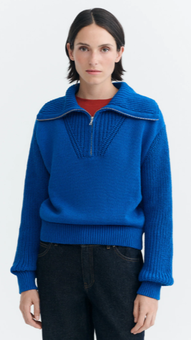 Woman wearing a bright blue sweater with a zippered collar, standing against a plain white background.