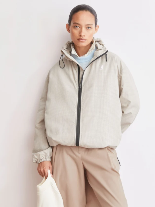 A woman wearing a light beige hooded jacket, tan pants, and holding a white bag, standing against a plain white wall.