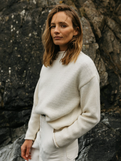 Woman with shoulder-length brown hair wearing a white sweater and white pants, standing outdoors in front of dark rocky terrain.