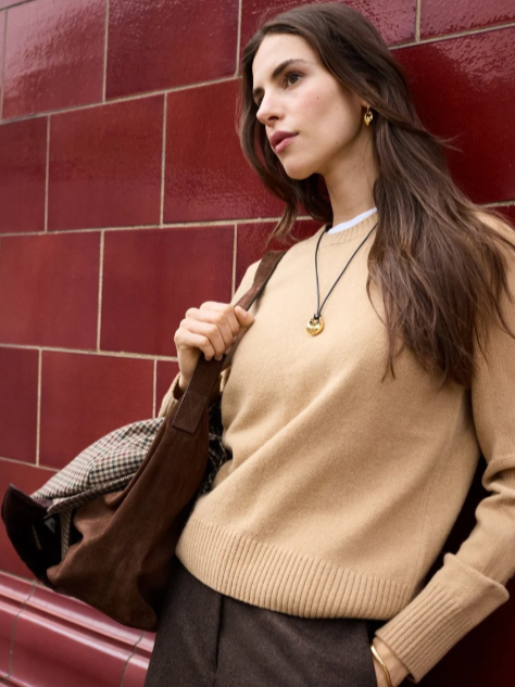 A young woman with long brown hair standing against a red brick wall, wearing a beige sweater, dark pants, gold jewelry, and carrying a brown bag over her shoulder.