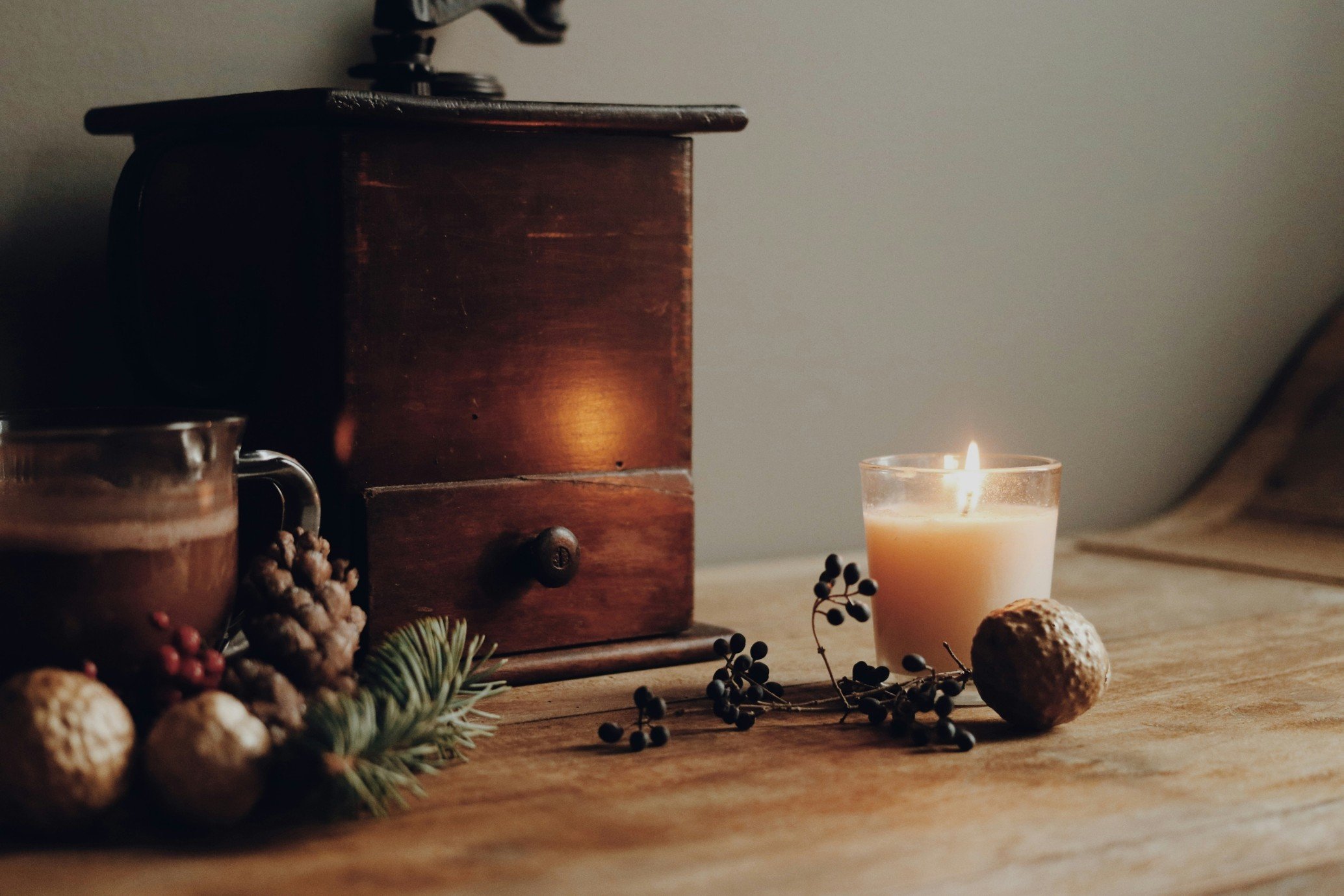 A cozy scene featuring a lit white candle, an antique coffee grinder, pinecones, berries, and decorative greenery on a wooden surface, with a plain wall in the background.