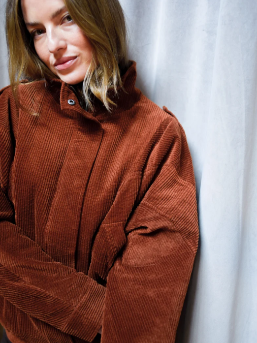 A woman with shoulder-length blonde hair wearing a brown corduroy jacket standing against a white curtain.