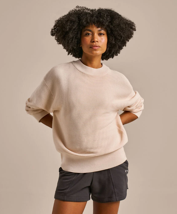 Woman with curly hair wearing a beige oversized sweater and dark shorts standing with hands behind her back against a neutral background.