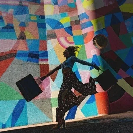Person dancing in front of a colorful geometric mural, holding two black bags and a cup.