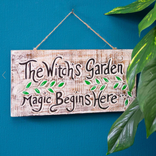 A wooden sign hanging on a blue wall that reads, "The Witch's Garden Magic Begins Here," with decorative green leaves and some large green plants partially visible on the right side.