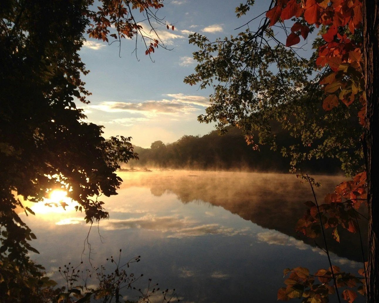 Sunrise or sunset over a calm river, with mist rising from the water and trees with autumn leaves framing the scene.