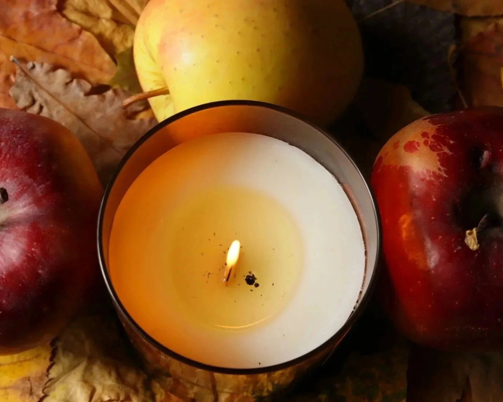 A lit white candle in a metallic holder surrounded by red and green apples, autumn leaves, and a yellow apple