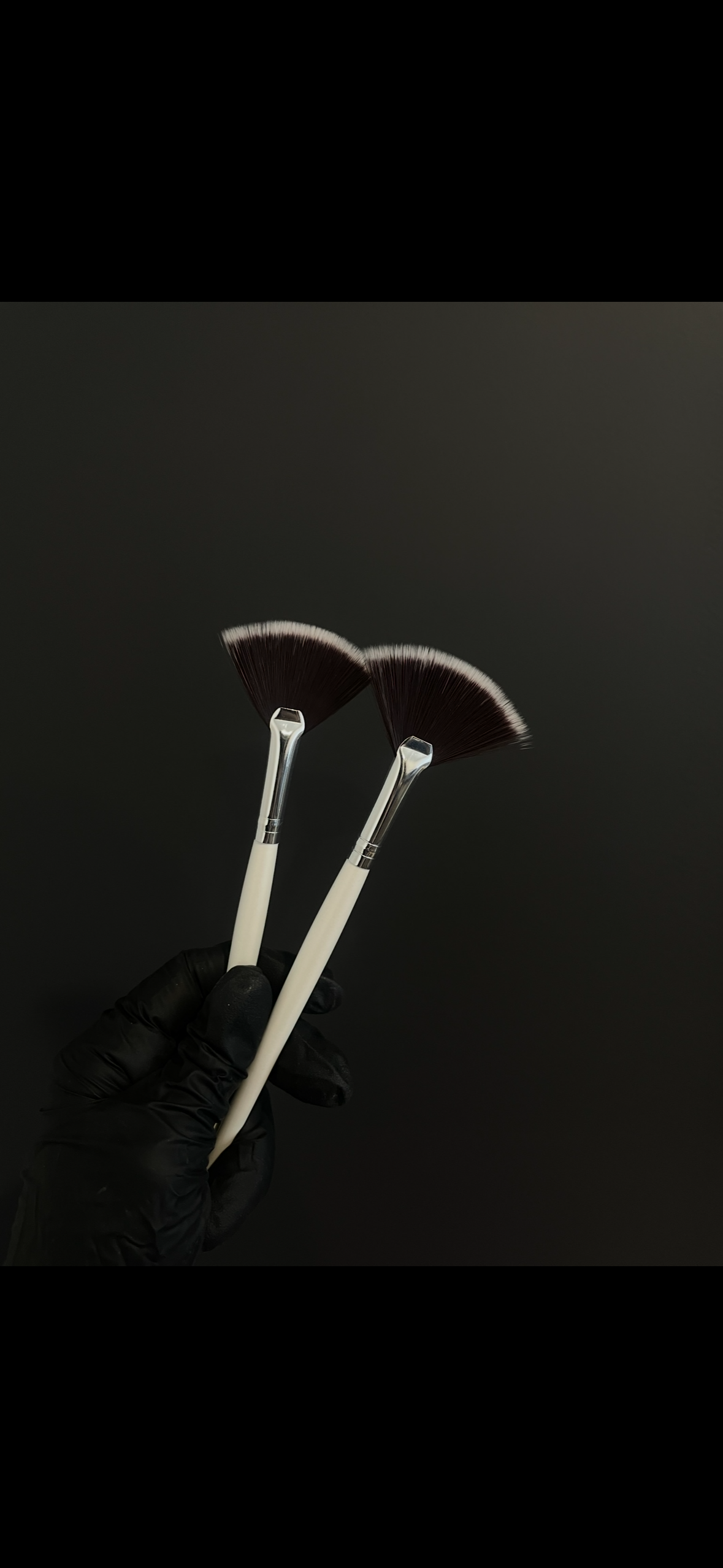 Two makeup brushes with fan-shaped bristles being held by a hand wearing a black glove against a dark background.