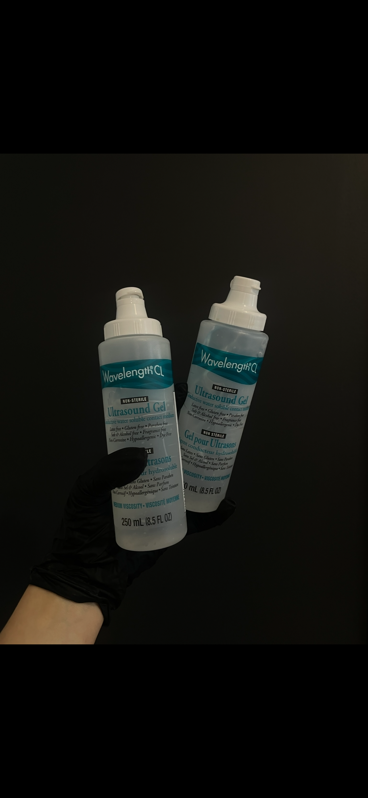 A person wearing a black glove holding two bottles of ultrasound gel against a dark background.