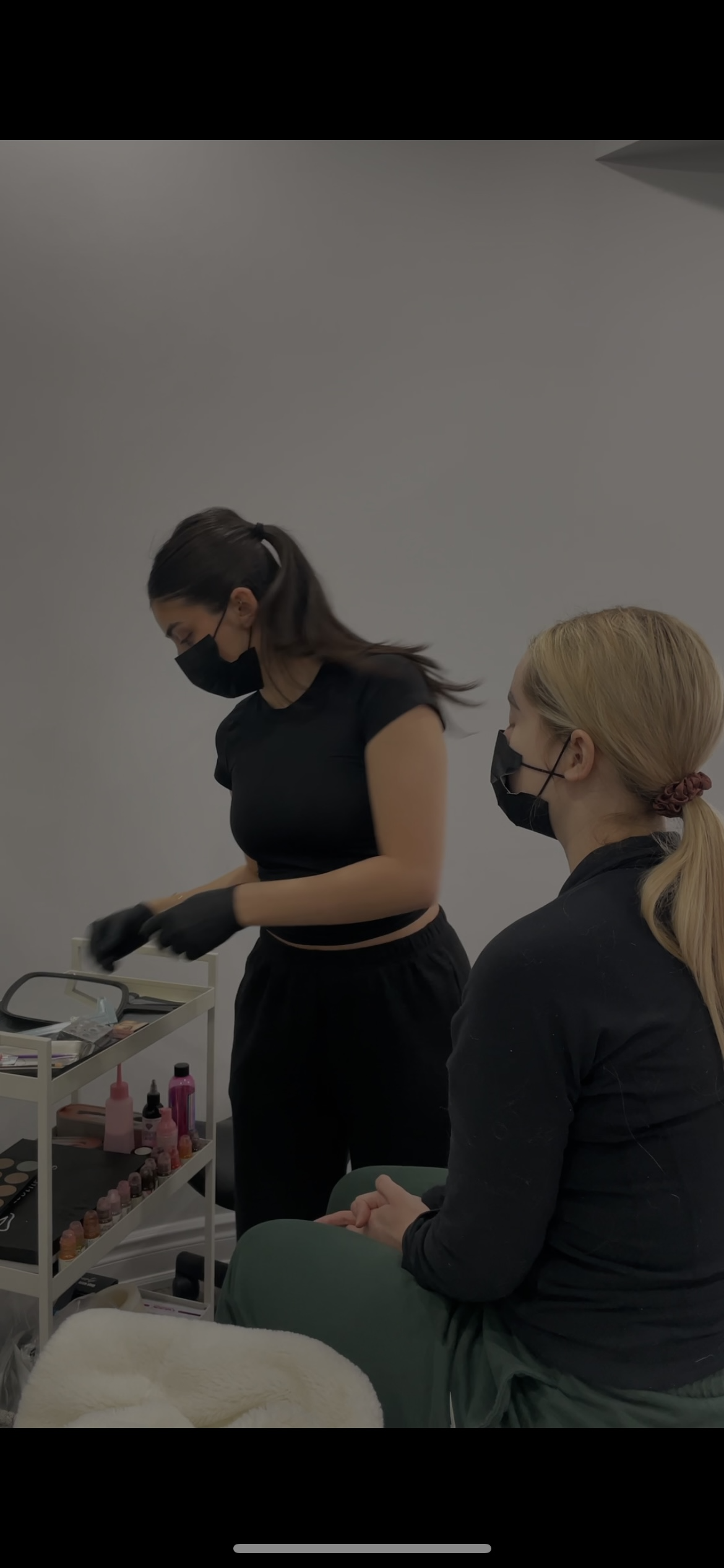 Two women in black shirts and masks, one sitting and one standing, wearing gloves and working with various beauty or skincare products on a cart.