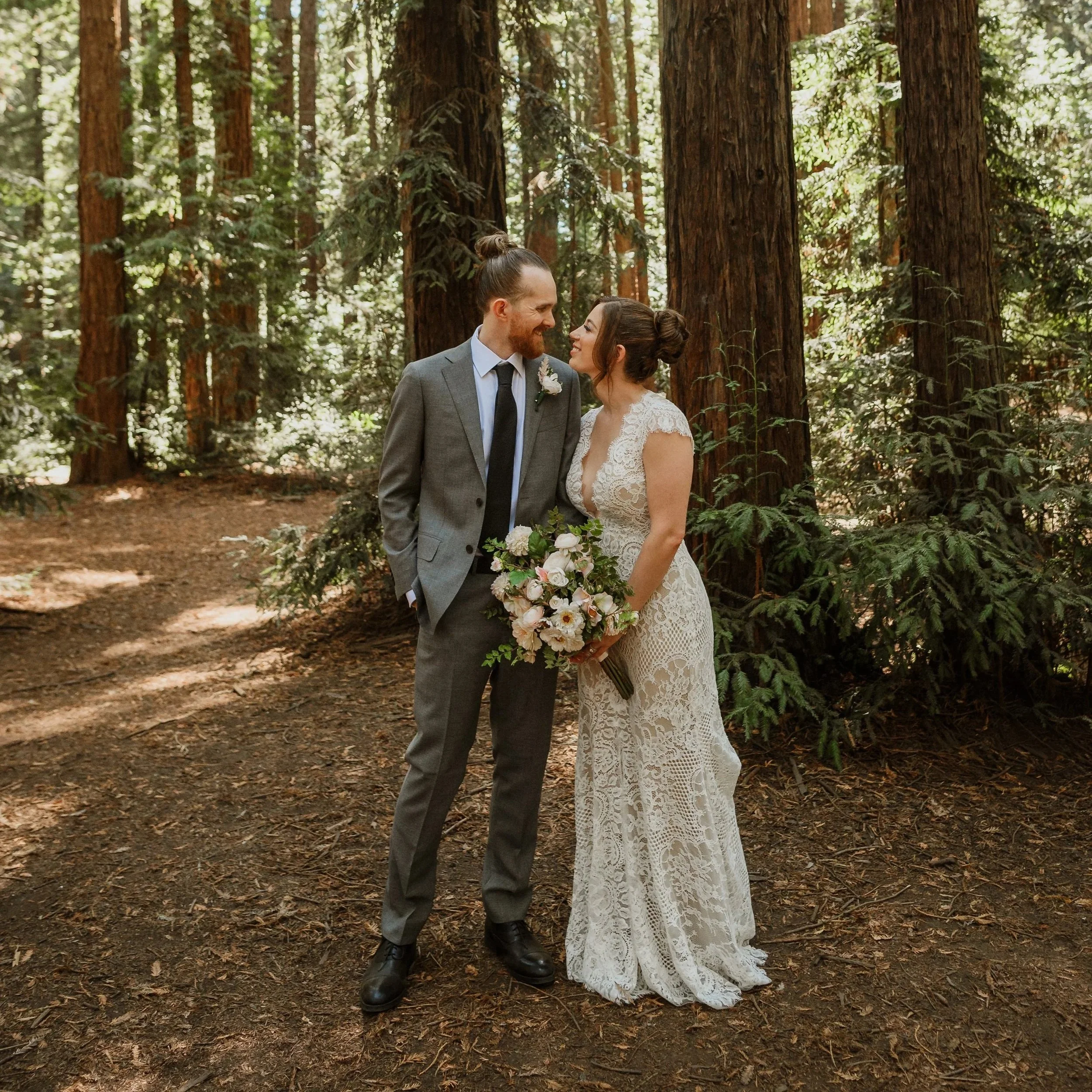 A bride and groom standing close together in a forest, smiling at each other. The groom is wearing a gray suit with a white shirt and black tie, and the bride is wearing a white lace dress and holding a bouquet of flowers. Tall trees surround them with sunlight filtering through the leaves.