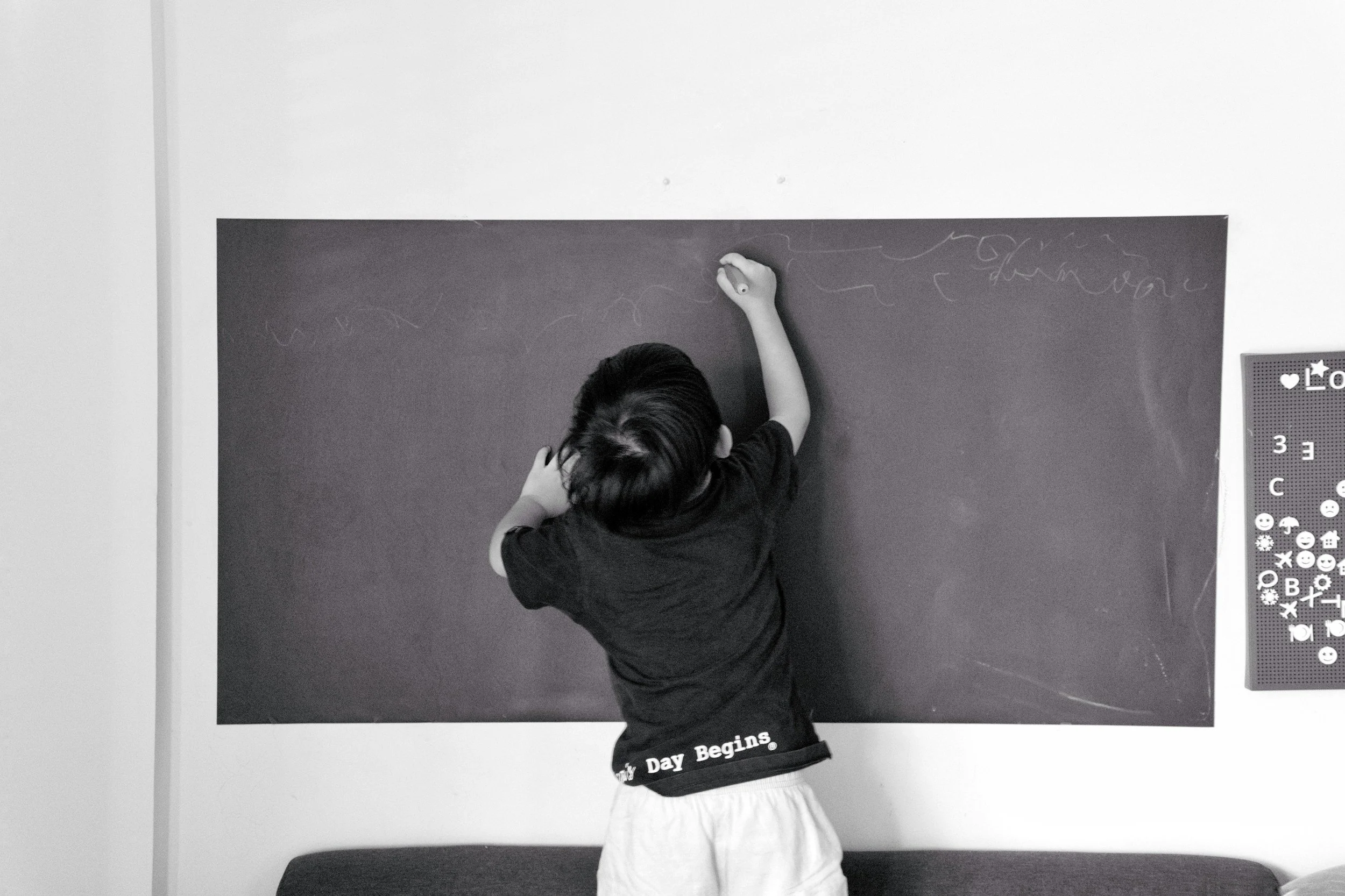 A child standing in front of a black chalkboard, drawing with chalk. The child is wearing a dark shirt and light-colored shorts, with the words 'My Day Begins' on the waistband. The photo is in black and white.