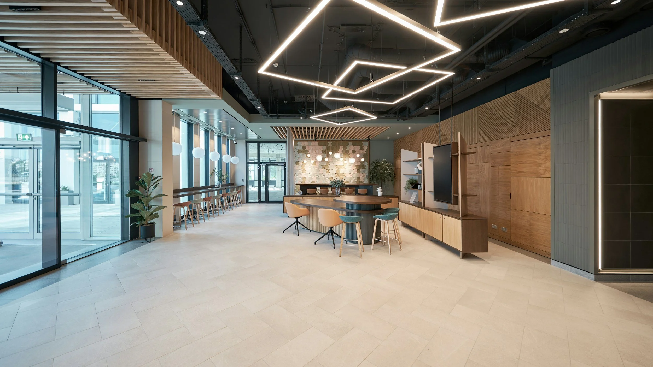 Modern indoor lobby with wooden accents, seating area, decorative lighting, large glass doors, and plants.