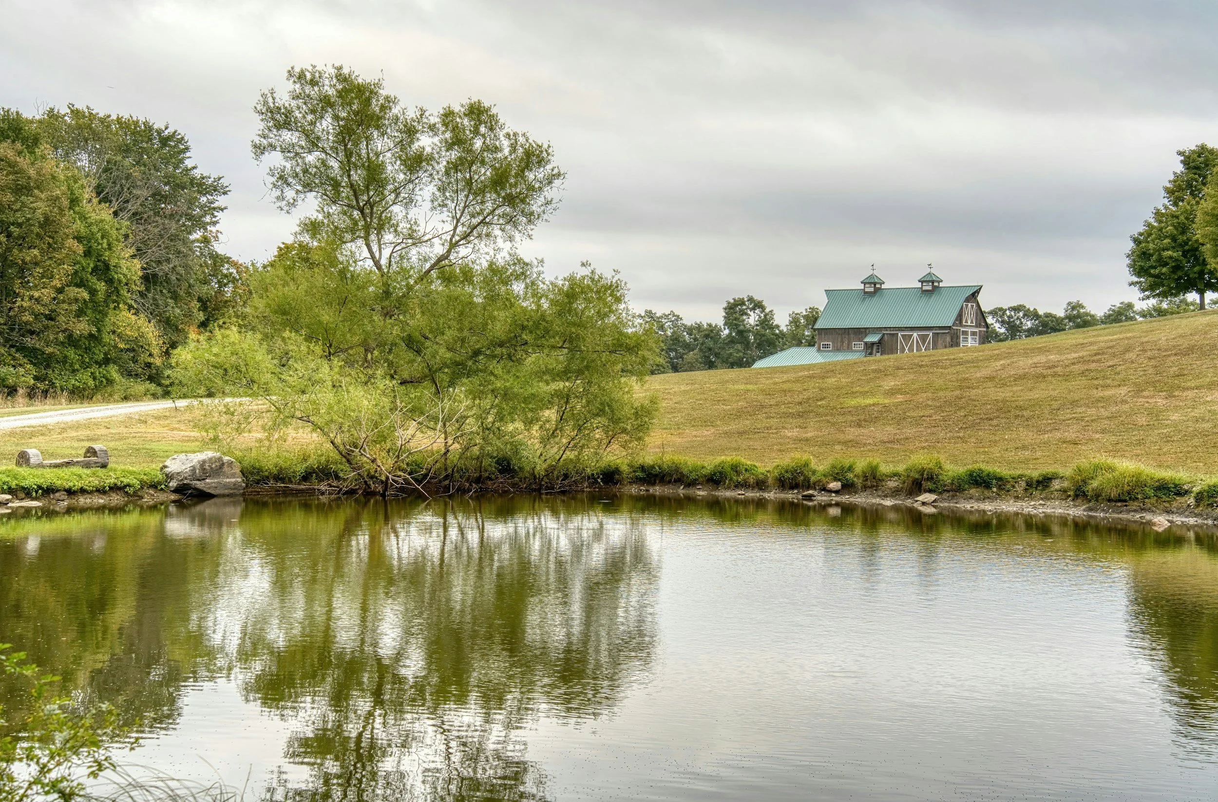 A scenic view of a pond with trees on the side and a barn on a hill in the background, under cloudy skies.