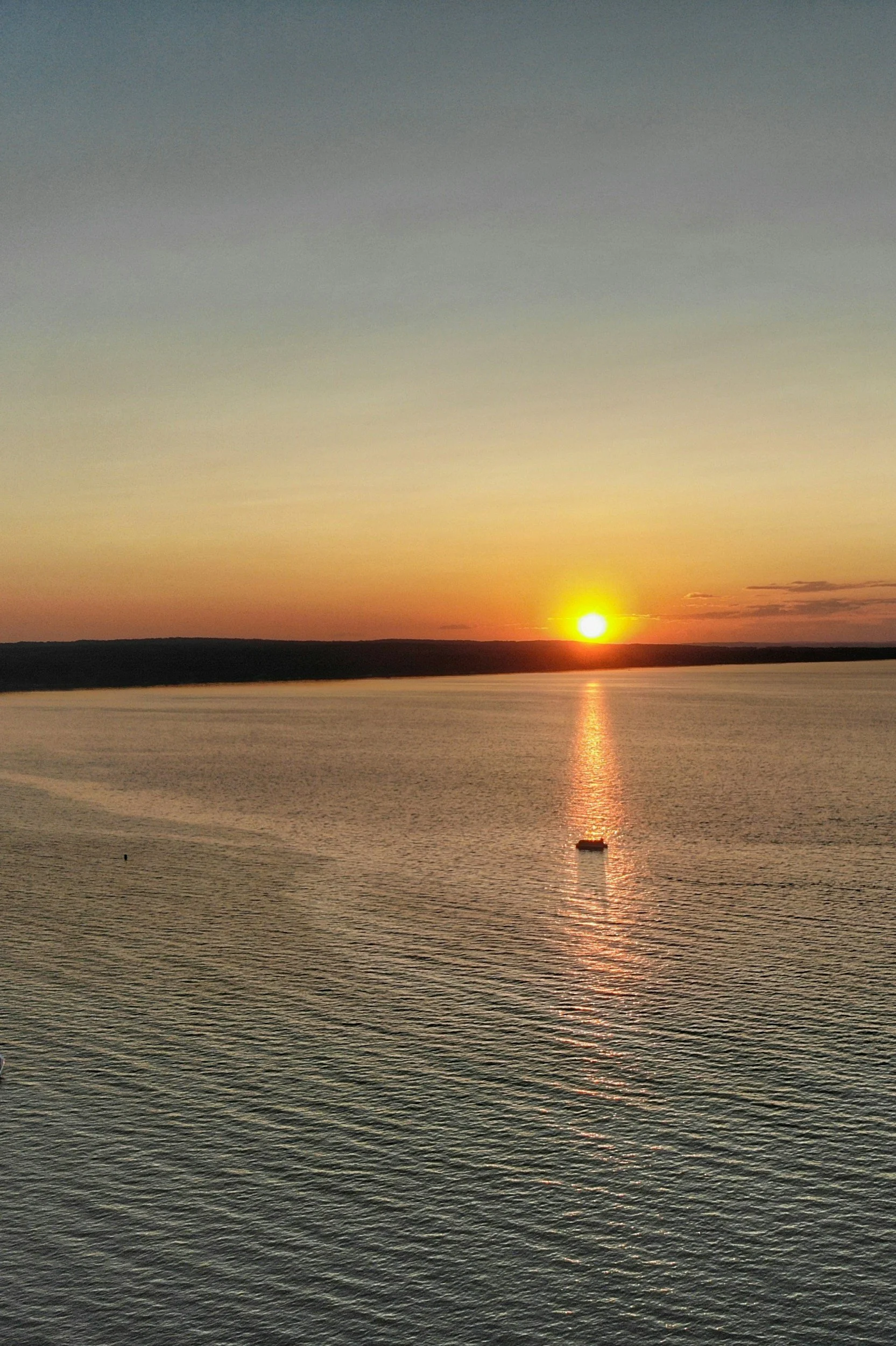 Sunset over a body of water with a small boat near the center and a dark shoreline in the background.