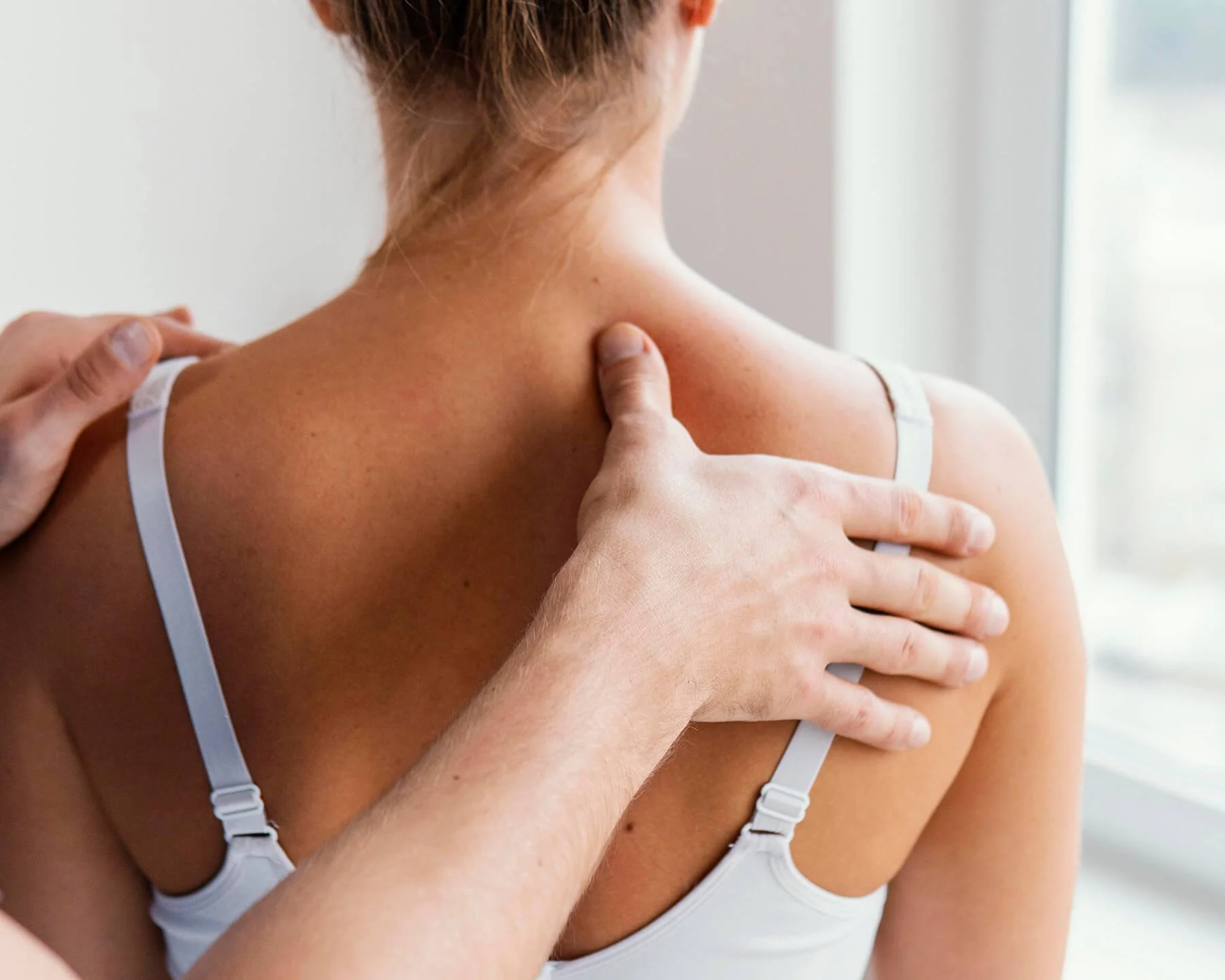 A person with light skin is holding their shoulder, which is being gently massaged or examined by another person. The individual is wearing a white strap top, and the background appears to be indoors with a window.