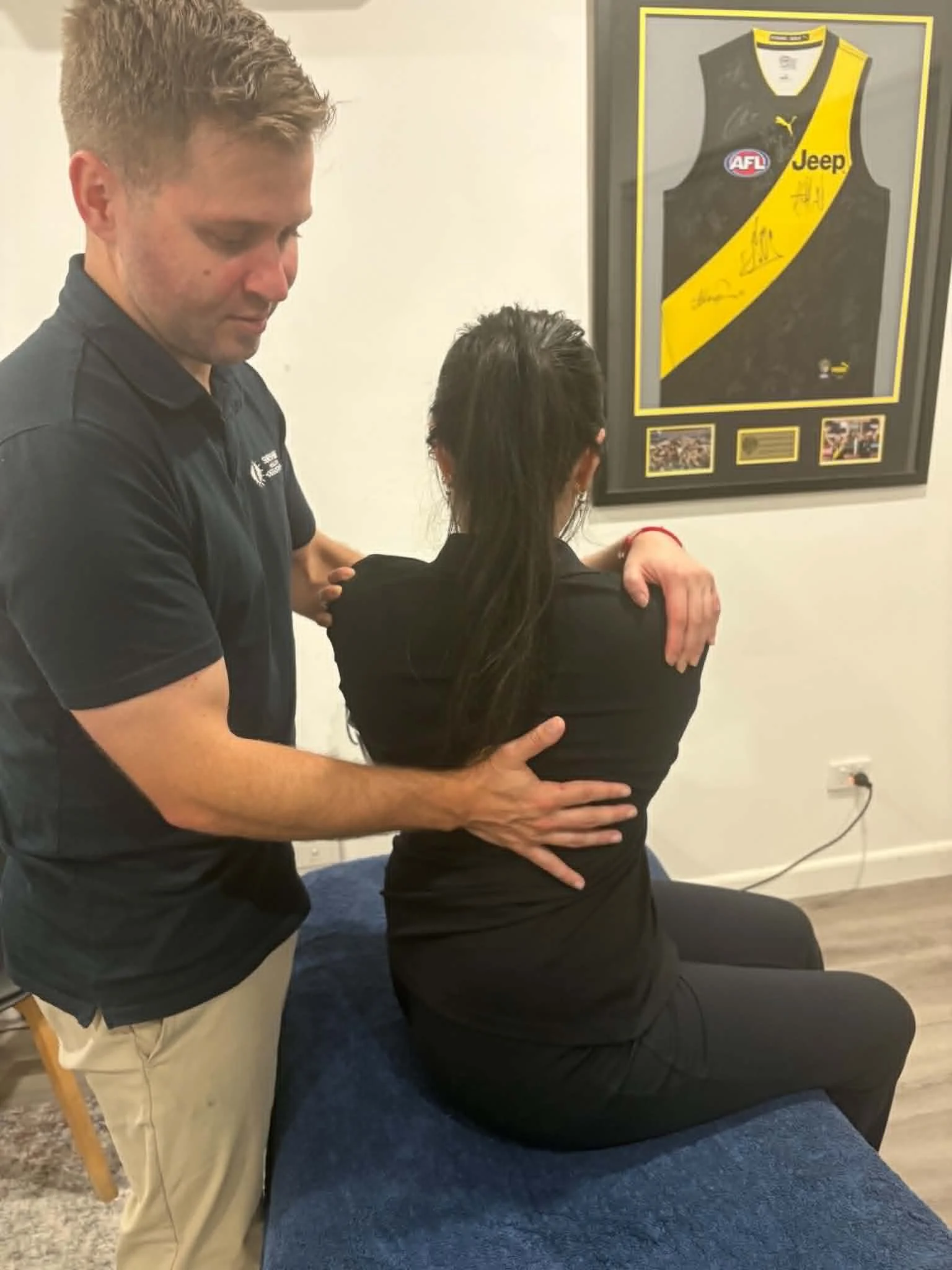 A man providing a chiropractic adjustment or massage to a woman seated on a blue upholstered table inside a room with a framed sports jersey on the wall