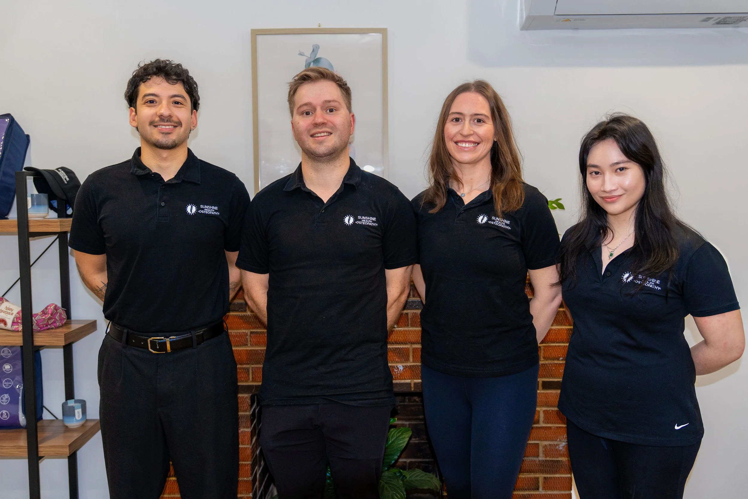Four people standing side by side, wearing matching black polo shirts with a 'Sunshine' logo, smiling in front of a brick and white wall with a framed picture and a plant in the background.