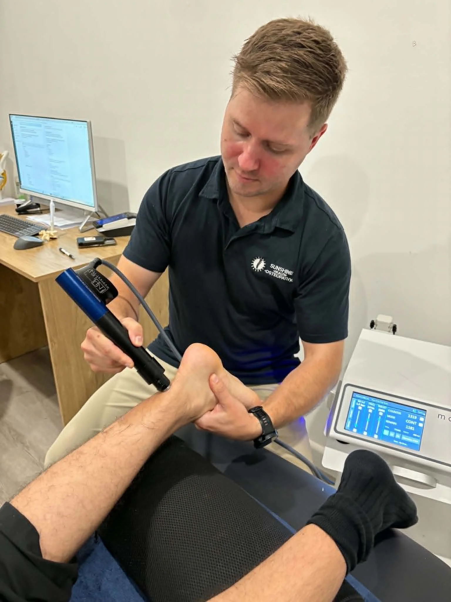 A healthcare professional performing a physical therapy treatment on a patient's arm using a handheld device, with monitoring equipment and a computer screen in the background.