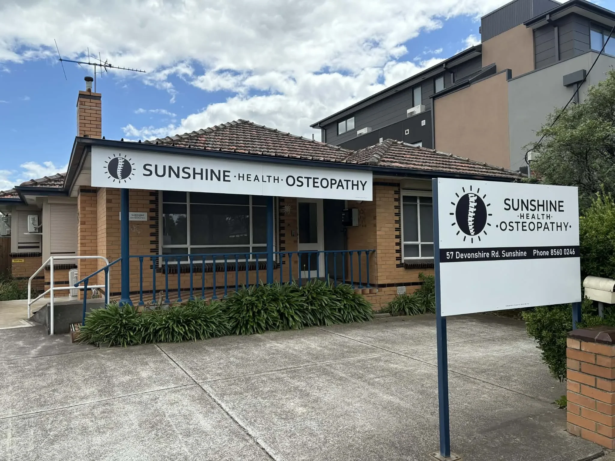 A building with a sign that reads 'Sunshine Health Osteopathy' and an address '57 Devonshire Rd. Sunshine' with a phone number. The building has brick walls, large windows, a red tiled roof, and a white and black sign with a logo that features a spine within a circle. There is a sidewalk with planted greenery in front of the building and a larger modern building in the background. The sky is partly cloudy with patches of blue.