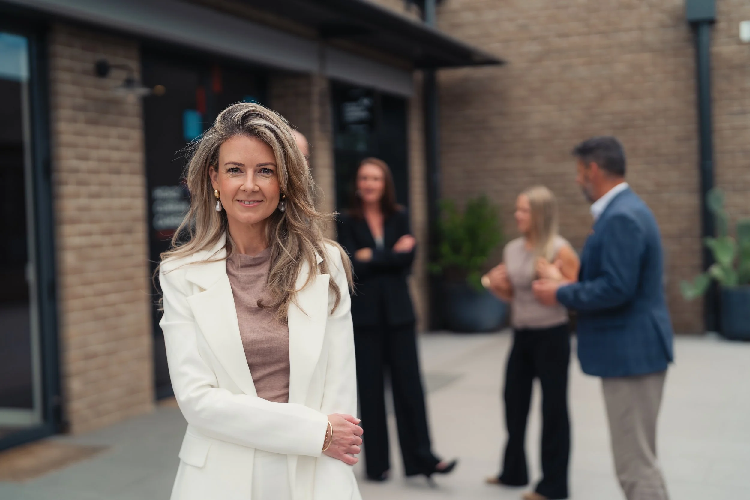 A woman with long, wavy blonde hair and wearing a white blazer with a beige top, smiling at the camera outdoors with a group of people in the background near a brick building.