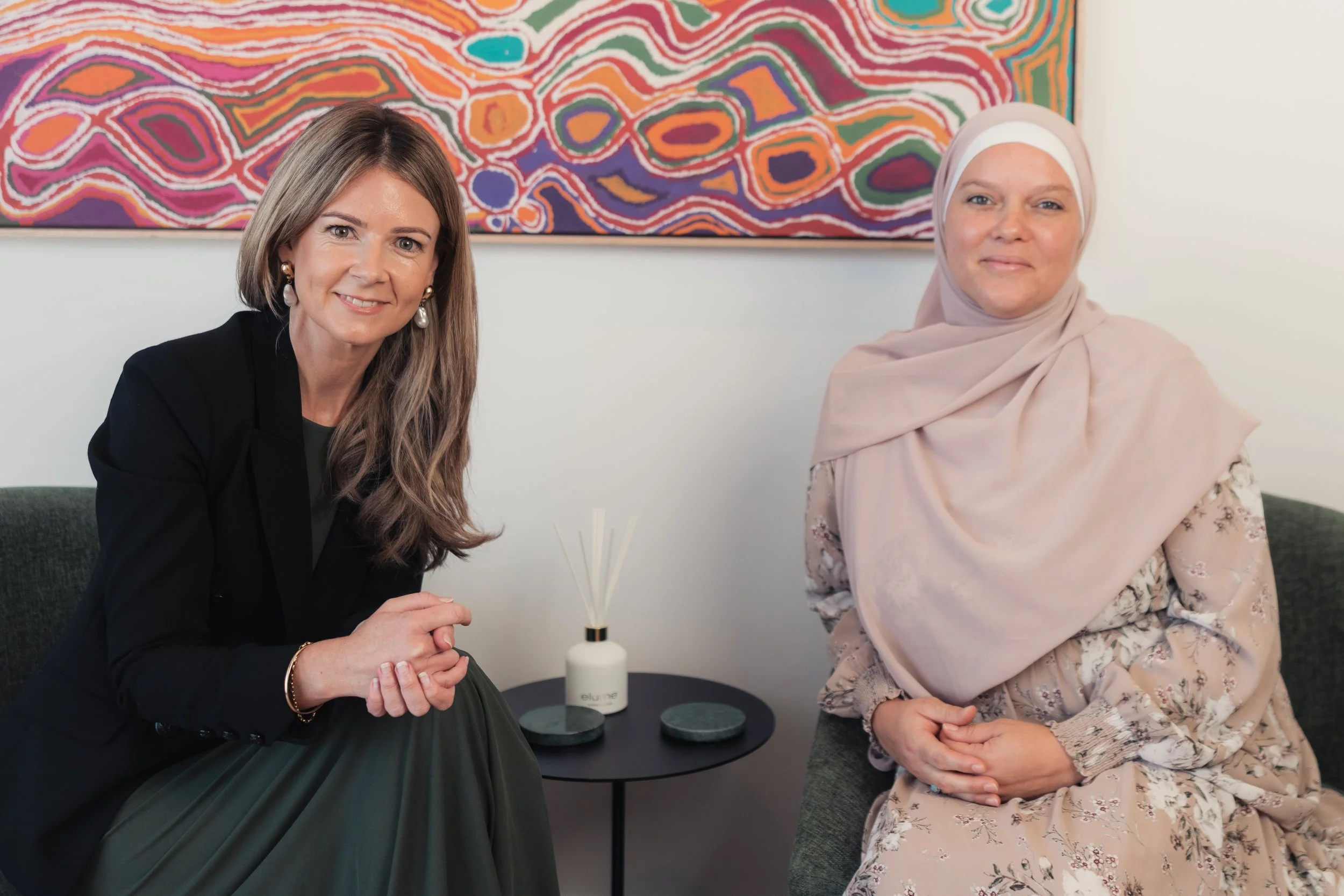 Two women sitting on a couch in front of a colorful abstract painting. The woman on the left is Caucasian, with long light brown hair, wearing a black blazer and pearl earrings, holding a white bottle with reed diffuser sticks. The woman on the right is Muslim, wearing a light-colored hijab and a floral dress, with her hands resting on her lap.