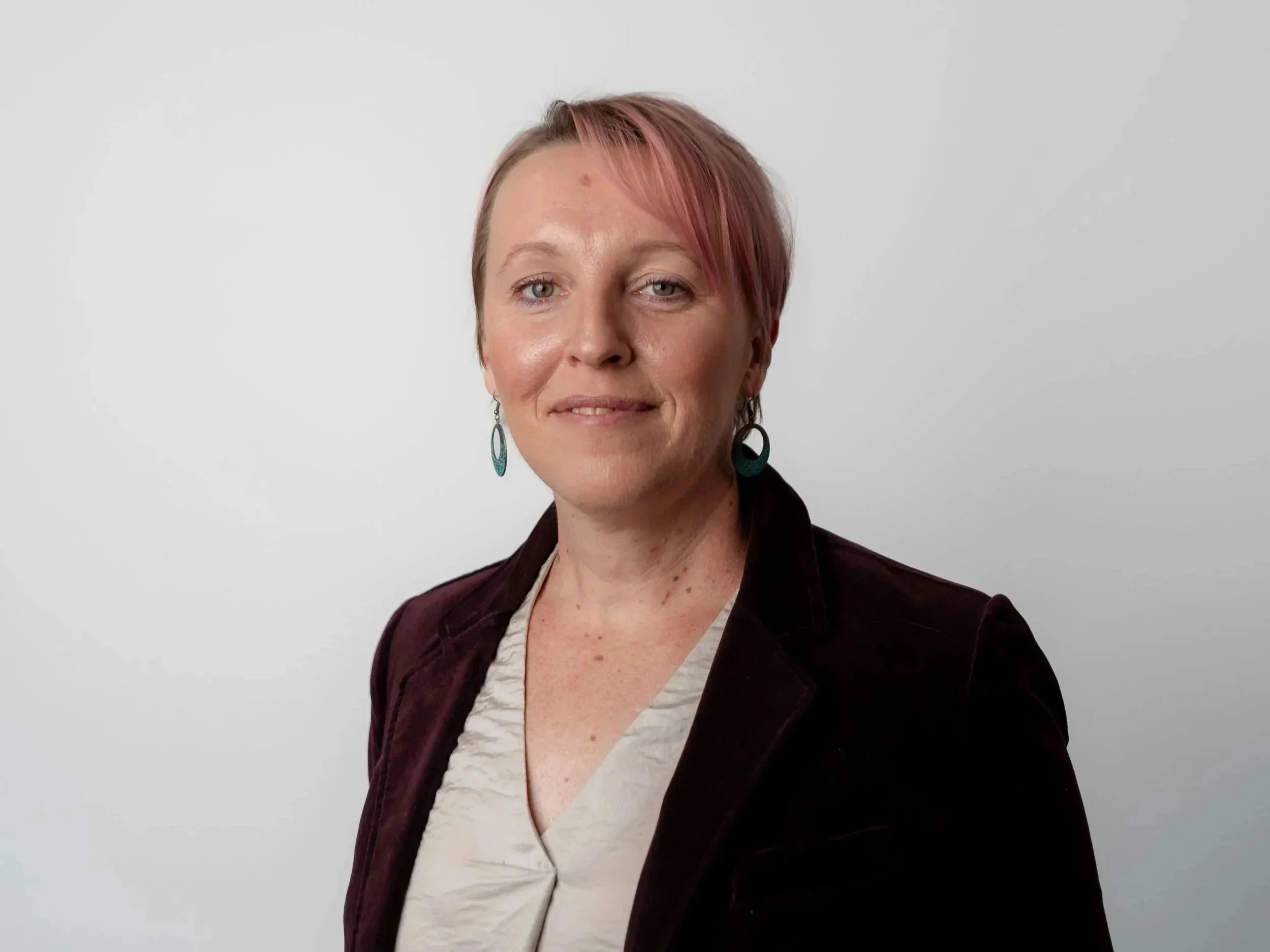 A woman with short, reddish-brown hair and earrings, wearing a jacket and a patterned shirt, standing against a plain light gray background.