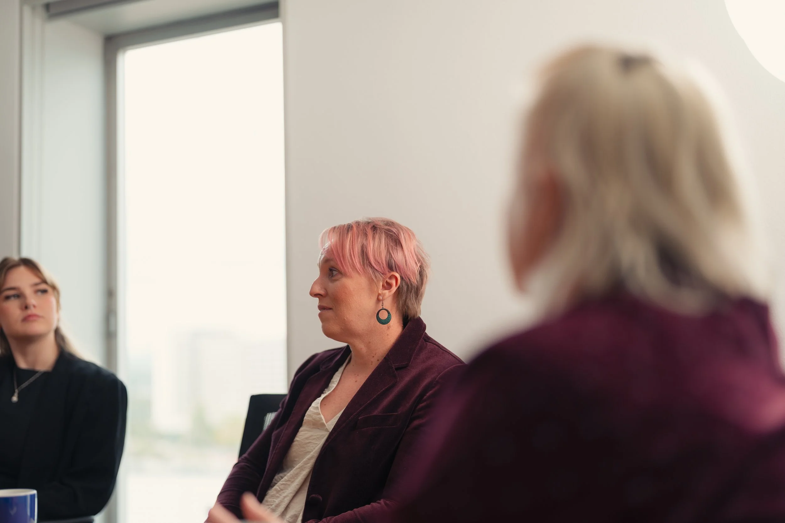 A group of people sitting at a table during a meeting in a bright room. The focus is on a woman with short pink hair and earrings, wearing a maroon blazer, looking to the side. In the foreground, part of another person's head with gray hair is visible. There is a window with blinds in the background.