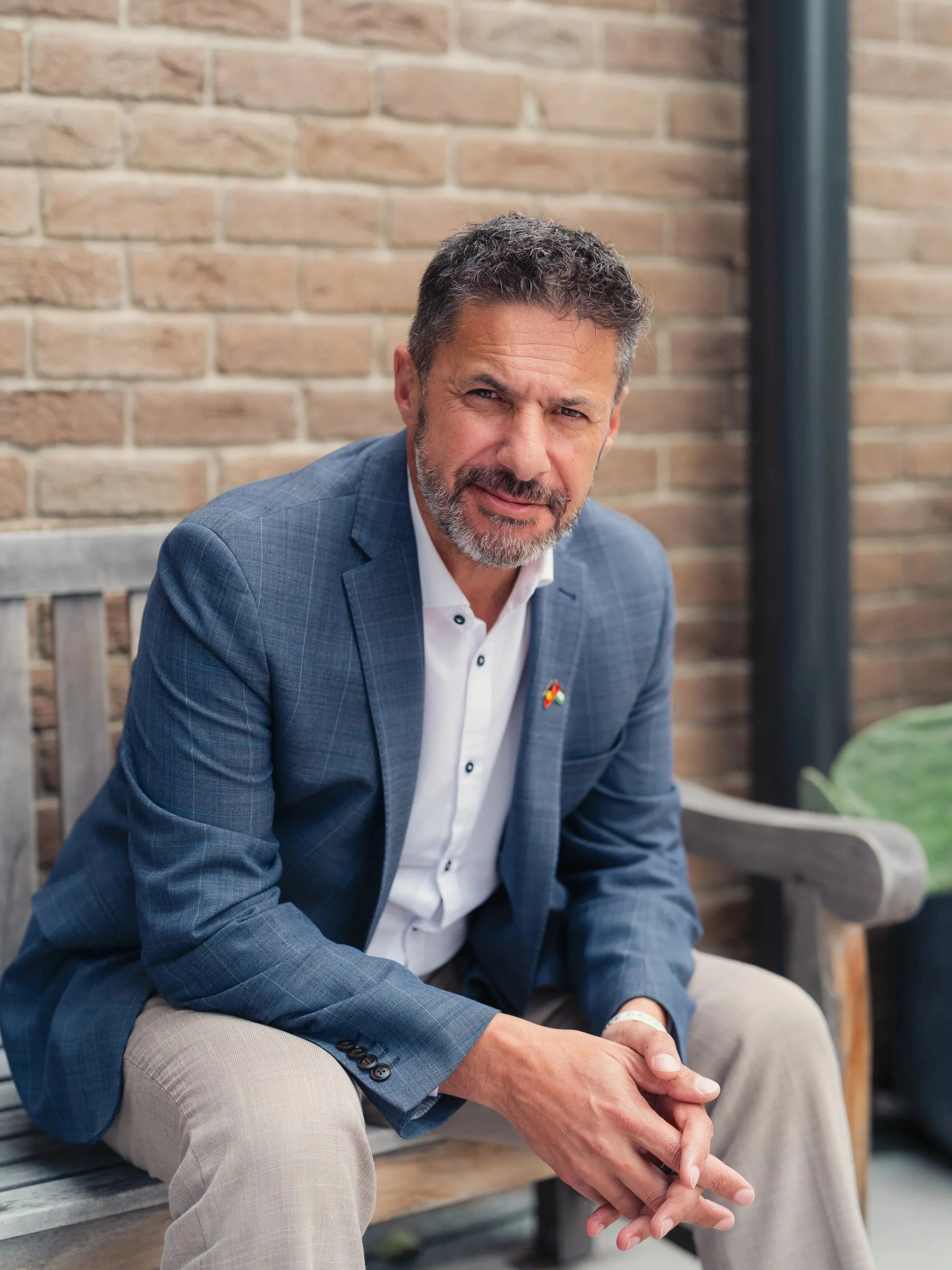 A middle-aged man with a beard, wearing a blue checked suit, light-colored pants, and a white shirt, is sitting on a wooden bench with a brick wall background. He has a small flag pin on his suit lapel.