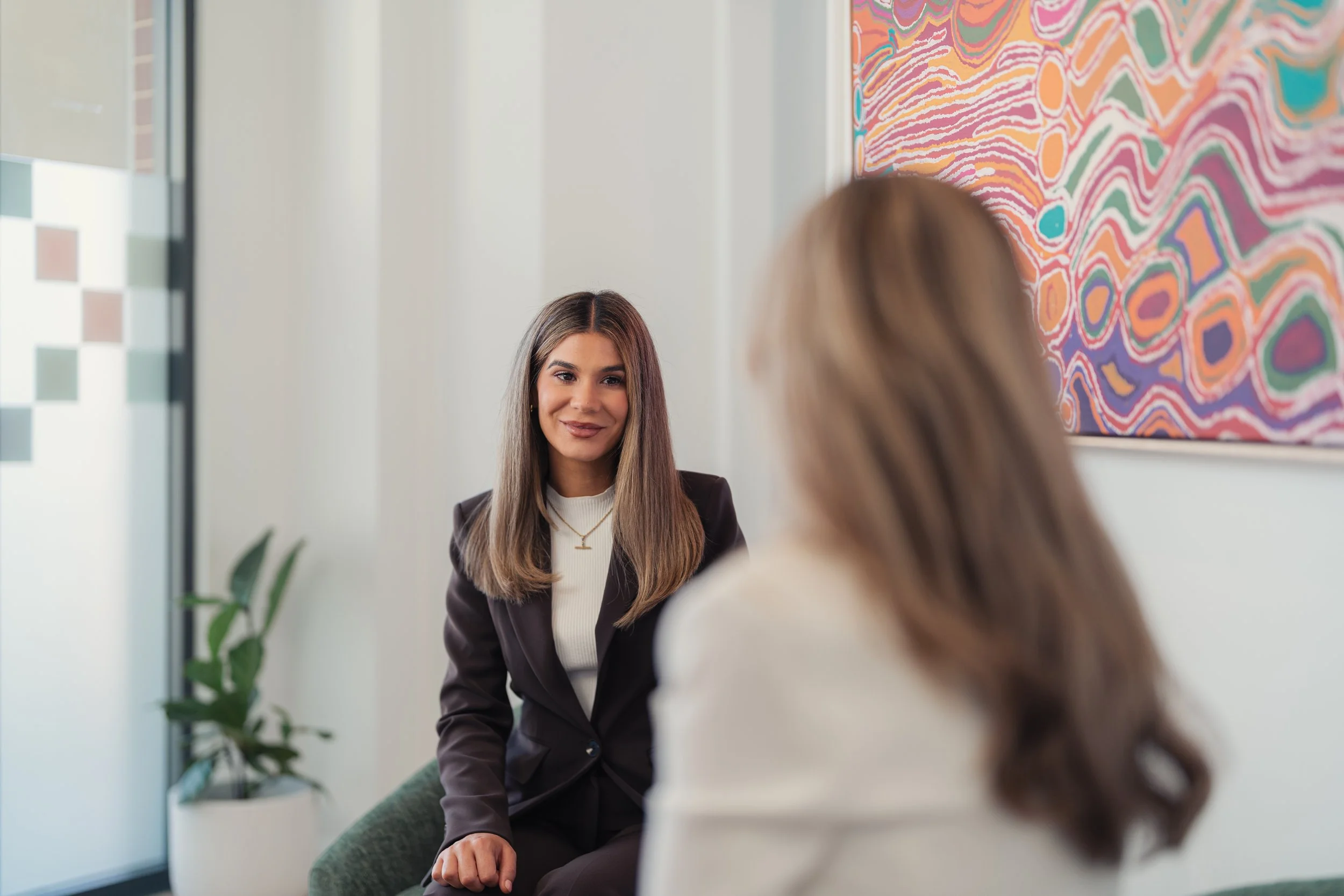 A young woman with light brown hair and a white shirt talking to another woman with long hair, wearing a dark blazer, in an office or gallery setting with an abstract colorful painting on the wall.