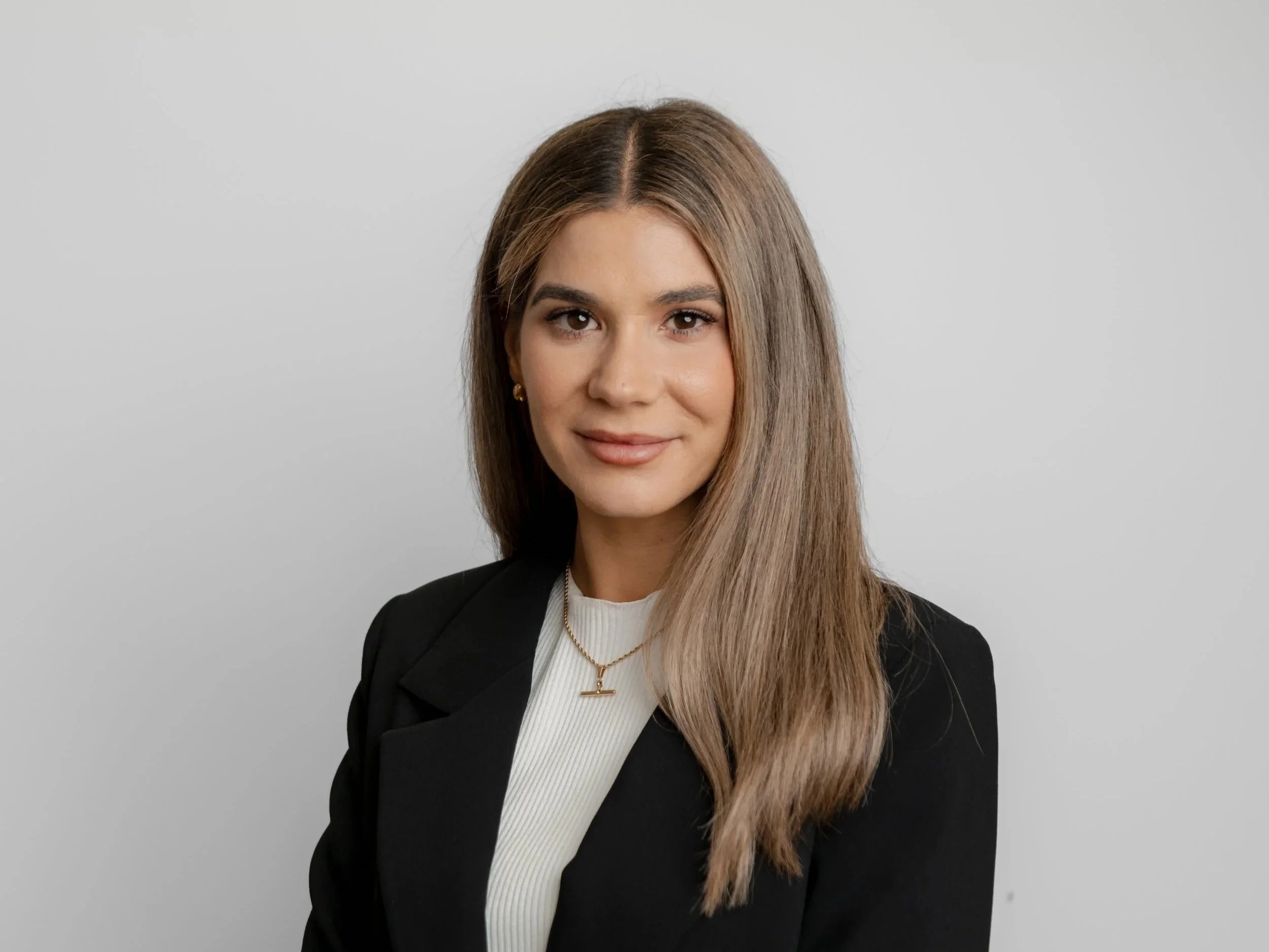 A woman with long light brown hair wearing a black blazer, white top, and gold jewelry smiling at the camera against a light gray background.