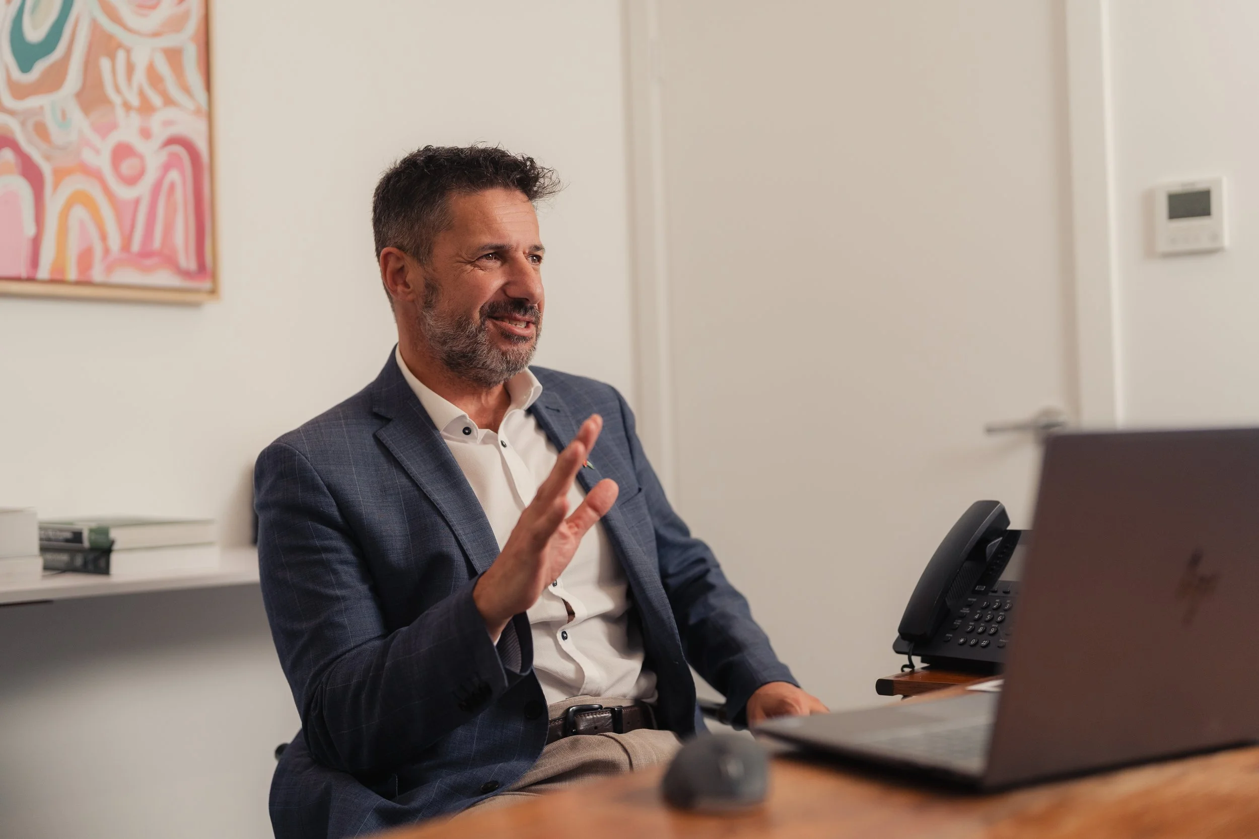 A middle-aged man with dark hair and a beard, dressed in a suit, sitting at a desk during a video conference, gesturing with his hand while speaking.