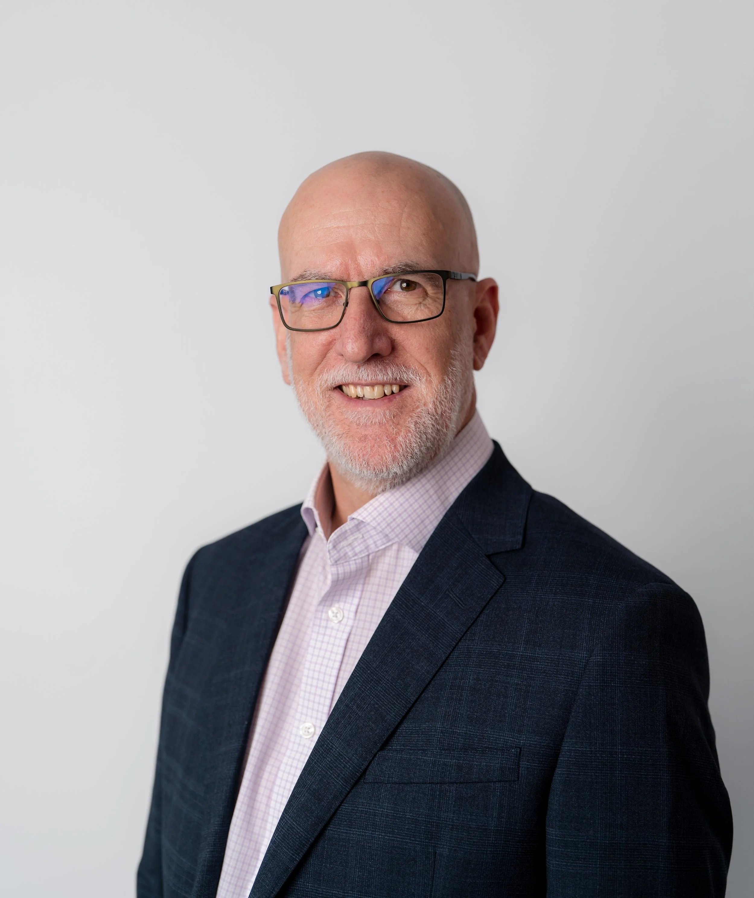 Portrait of an older man with a bald head, glasses, a white beard, wearing a dark blue suit and light pink checkered shirt, smiling, in front of a plain light gray background.