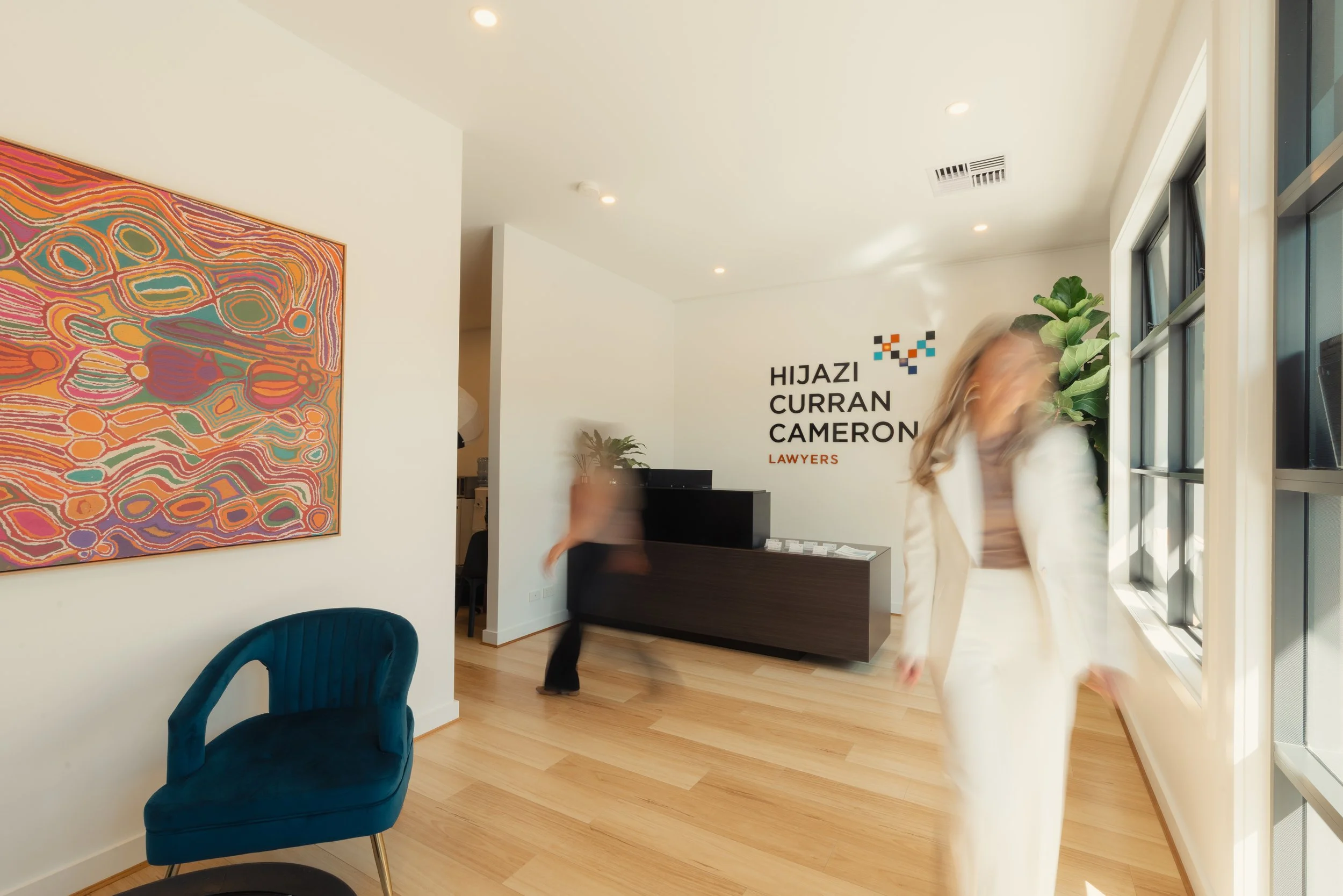 Reception area of law office with colorful abstract art on the wall and a sign that reads 'HIJAZI CURRAN CAMERON LAWYERS.' There is a teal velvet chair in the foreground, a woman walking past blurred, and a potted plant near the large windows.