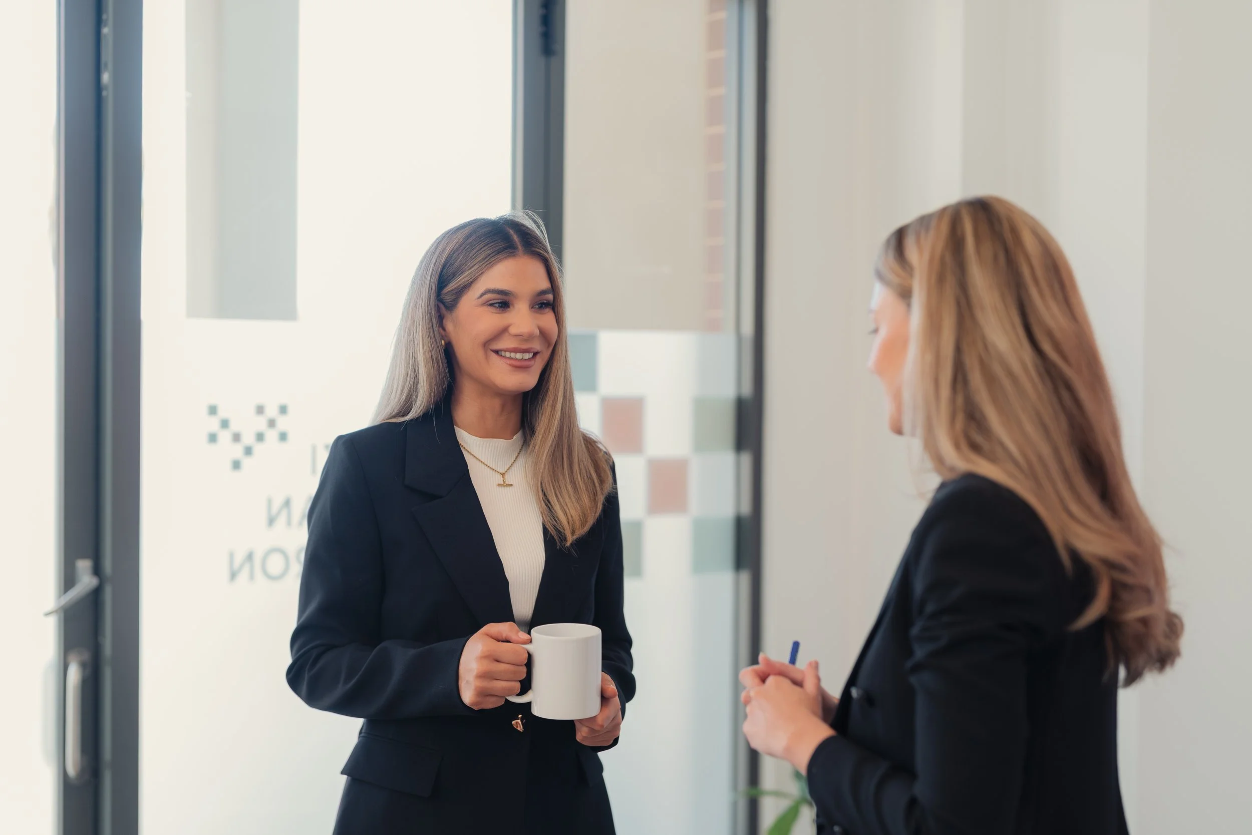 Two women having a conversation in an office, one holding a white mug.
