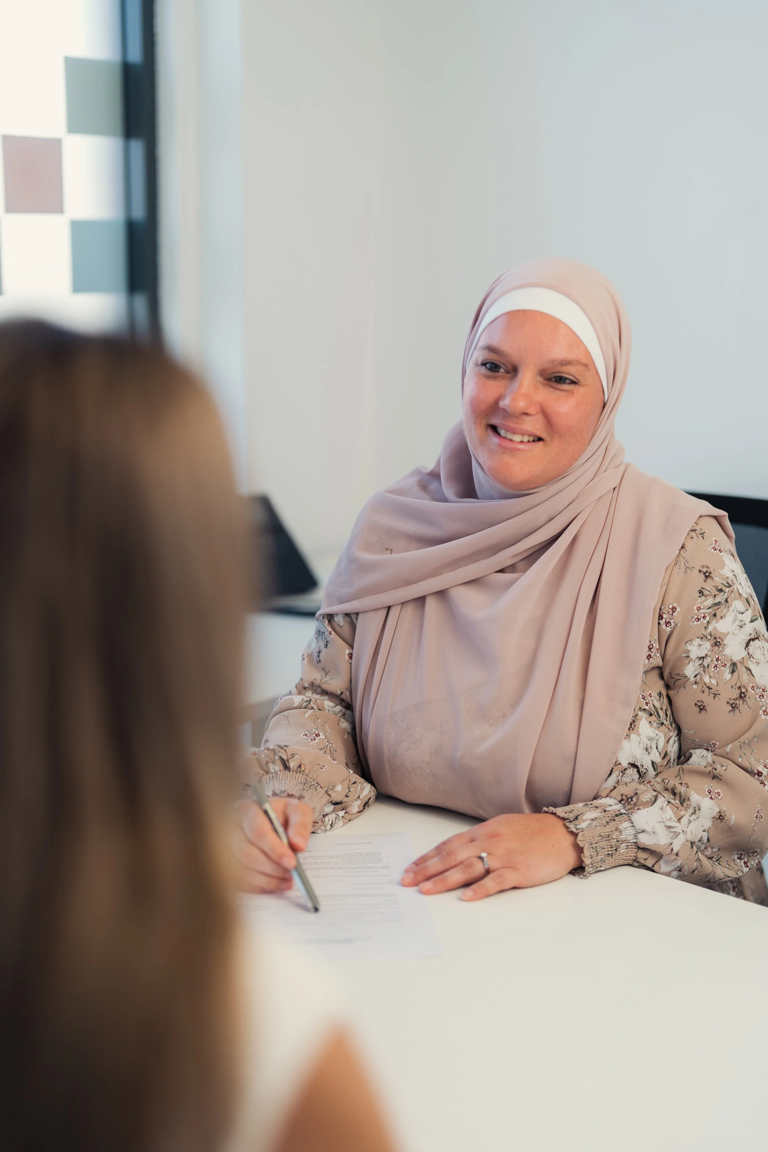 A woman wearing a beige headscarf and floral blouse smiling while sitting at a table during a discussion or interview.